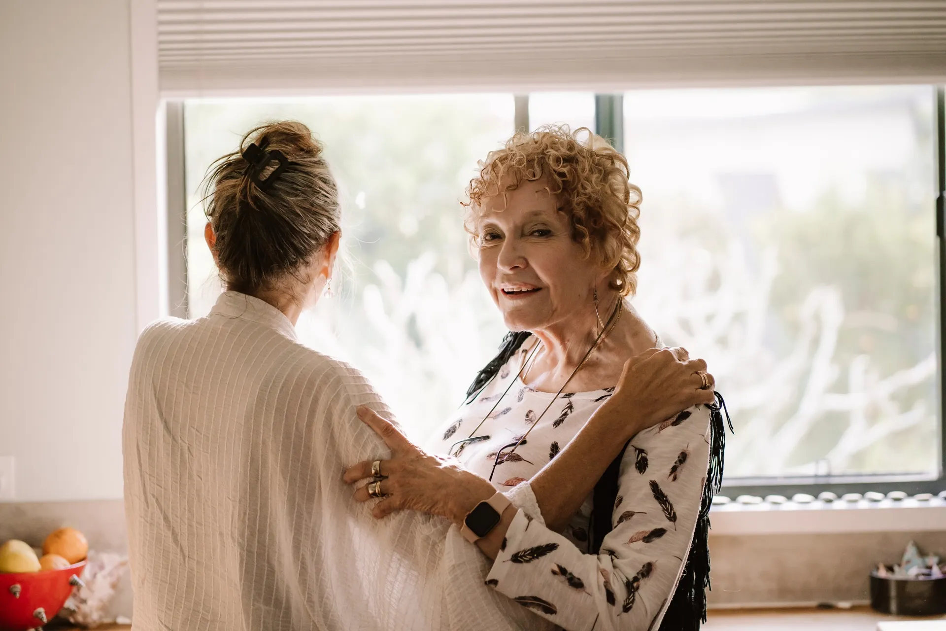 An older woman and a carer embracing warmly by a sunlit window at home